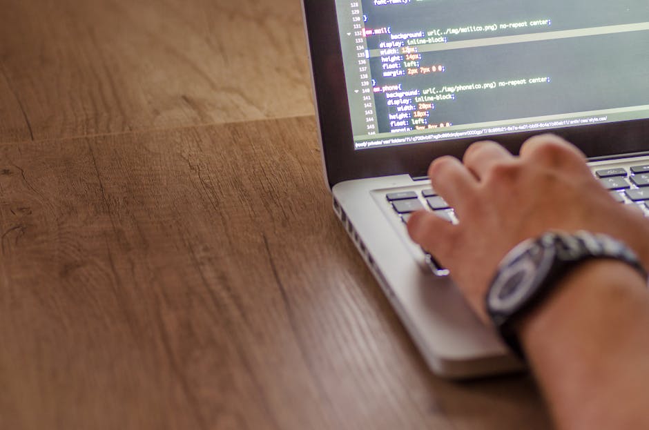 The image shows a person's hand typing on a laptop keyboard placed on a wooden desk. The laptop screen displays lines of code or a website's HTML, with colors indicating different syntax elements. The
