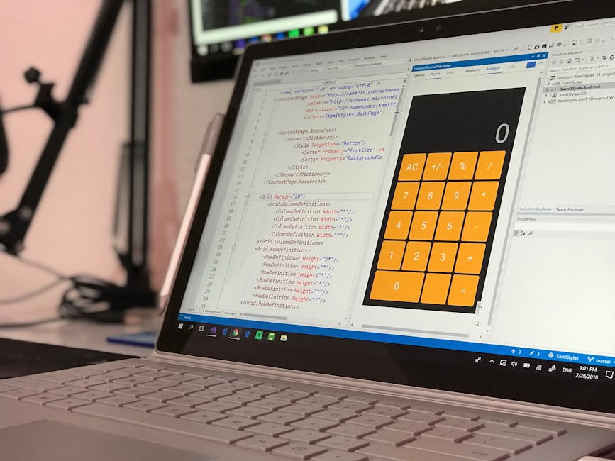 A close-up view of a laptop screen displaying lines of coding and a large, orange calculator app with basic arithmetic functions, set on a desk in a workspace. The background features blurred equipmen